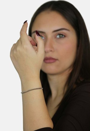 Bracciale d'argento indossato su un polso, rotondo e liscio, con una finitura fine. La mano è sollevata, mostrando il bracciale su uno sfondo neutro.