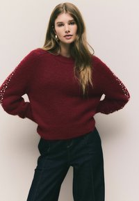 Burgunderfärgad stickad tröja med avslappnad passform, prydd med pärlaccent på ärmarna, i kombination med mörk denimjeans. Mjuk textur och rundad halsringning.