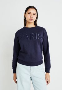 Sweatshirt azul-marinho com a inscrição "PARIS" em letras bordadas, decote redondo, punhos e bainha canelados, combinado com uns jeans azul-claro.