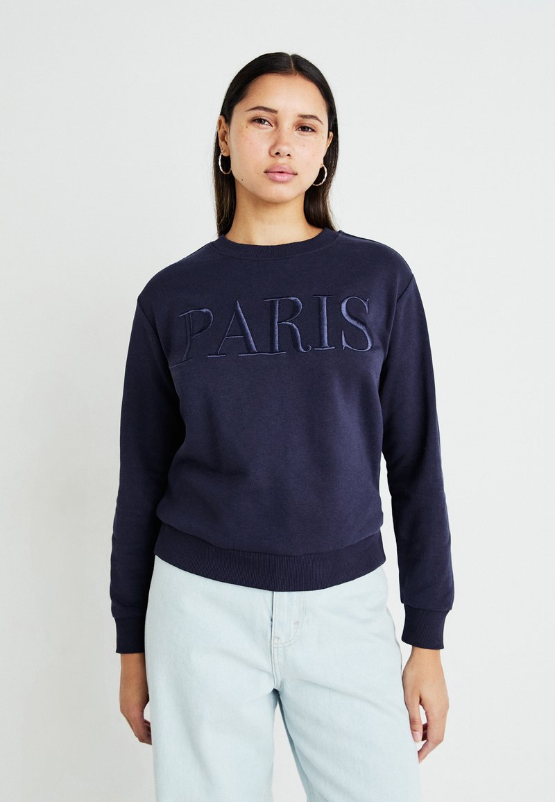 Sweatshirt azul-marinho com a inscrição "PARIS" em letras bordadas, decote redondo, punhos e bainha canelados, combinado com uns jeans azul-claro.