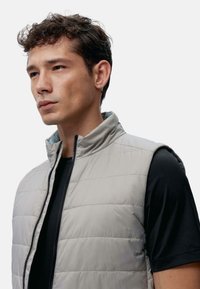 Lys grå puffer vest med høy krage, vertikal søm og glidelås foran. Brukt over en svart t-skjorte med korte ermer.