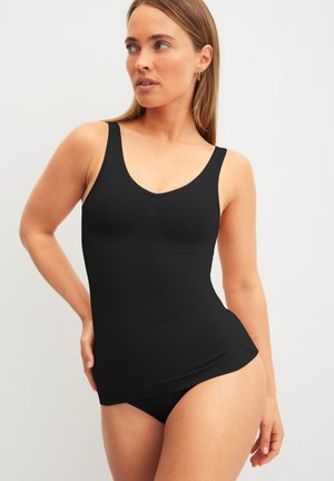 Frau trägt ein eng anliegendes schwarzes Tanktop und passende schwarze Unterwäsche, posiert mit einer Hand in der Hüfte und schaut seitlich.