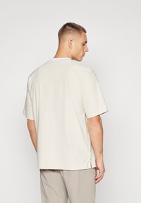 Tricou relaxat, cu mânecă scurtă, de culoare bej deschis, cu guler rotund și slituri pe laterale la marginile de jos. Textură de țesătură fină.