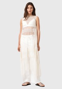 Durchsichtiges weißes Spitzenkleid mit mehrlagigem Chiffonrock, V-Ausschnitt und zarten Saumakzenten; kombiniert mit flachen, metallischen Sandalen.