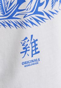 Camiseta de tela blanca con un diseño gráfico y texto en azul intenso, que incluye "ORIGINALS WORLDWIDE" en una tipografía moderna. Textura suave.