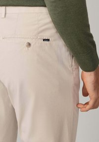 Bež chinos hlače s glatkom teksturom, s gumbom za zatvaranje i stražnjim džepom. Brand oznaka "HACKETT" vidljiva je na džepu.