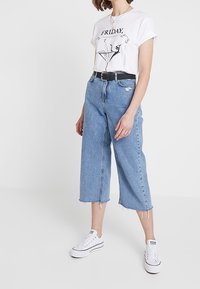 T-shirt bianca con una grafica nera che recita "VENERDÌ", abbinata a jeans cropped azzurri e sneakers bianche. Una cintura nera mette in risalto il punto vita.