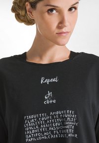 Schwarzes, kurzärmeliges Shirt mit weitem Kragen. Mit weißem Text: "Wiederholen" und verschiedenen tanzbezogenen Wörtern in einer verspielten Schriftart.