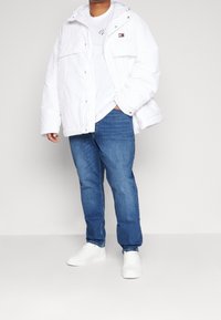 Hvid puffjakke med hætte, med knaplukninger og to forreste lommer, båret over en hvid t-shirt og blå jeans, komplet med hvide sneakers.