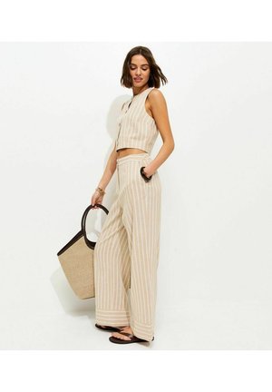 Vrouw in beige-wit gestreept mouwloos topje en brede broek die een grote strooien tas met donkere handvatten vasthoudt.