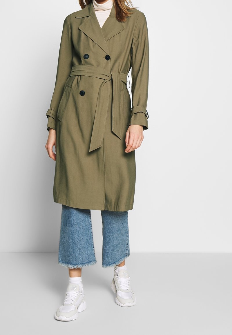 Kobieta w oliwkowym płaszczu typu trencz z paskiem, przedłużonych niebieskich dżinsach z postrzępionymi brzegami, białych skarpetkach i białych trampkach na jednolitym tle.