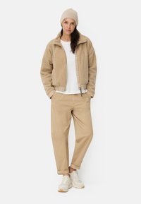 Veste bomber en polaire beige avec fermeture éclair, associée à un pantalon chino beige foncé et des baskets blanches. Le mannequin porte un bonnet beige clair. Design simple.