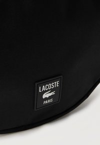 Mustast kangast pind, millel on ruudukujuline Lacoste Paris logo plaastriga, millel on valge krokodilliembleem.