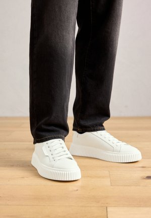 Persona che indossa sneaker bianche a basso profilo con suole spesse e jeans neri, in piedi su un pavimento in legno chiaro.