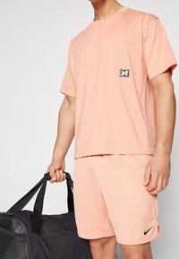 Men's sportswear set featuring a light peach mesh T-shirt with a small logo patch and matching shorts, holding a black duffel bag.