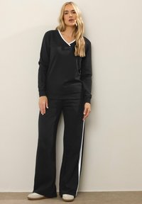 Sudadera negra con un ribete blanco en el cuello en V, combinada con pantalones negros que tienen rayas laterales blancas, ambos fabricados con tela suave. Calzado casual completa el conjunto.