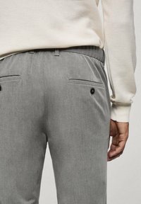 Homme portant un pantalon gris clair avec des poches arrière, habillé d'un pull blanc à manches longues texturé, main détendue le long du corps.