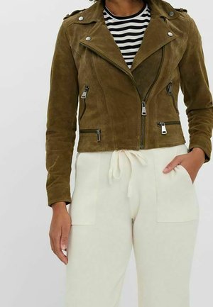 Vrouw draagt een olijfgroene suède ritssluiting jas over een zwart-wit gestreept shirt met crèmekleurige broek met koord, hand in de zak.