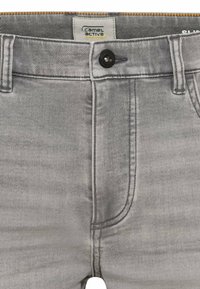 Graue Jeans aus Denim mit schmaler Passform, die über einen frontalen Knopfverschluss, Gürtelschlaufen und typisches Taschen-Design verfügen. Das Etikett trägt die Aufschrift "Camel Active."