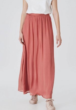 Femme portant un haut blanc, une jupe longue fluide couleur rouille et des baskets rose clair, debout devant un fond uni.