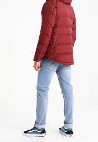 Röd pufferjacka med huva, strukturerad quiltning, blå denimjeans och svart-vita sneakers med blå accenter. Positionerad bakifrån.