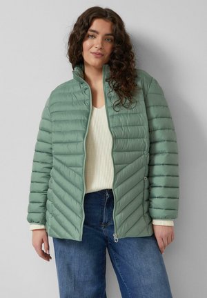 Frau mit lockigem Haar, die eine hellgrüne gesteppte Jacke über einem cremefarbenen Pullover und blauen Jeans trägt, steht vor einer grauen Wand.