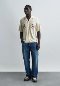 Hombre vistiendo una camisa de manga corta color crema con bordado de caballo rojo, jeans azules y zapatos multicolor, de pie con las manos en los bolsillos.