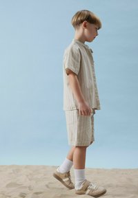 Camicia a maniche corte beige chiaro a righe e pantaloni corti coordinati, realizzati in un tessuto morbido. Sneakers marroni e calzini bianchi completano l'outfit.