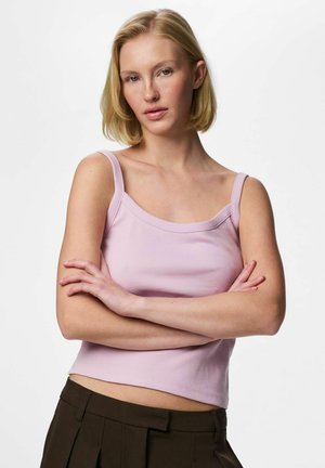 Fiatal szőke hajú nő, világos rózsaszín ujjatlan felsőt és sötét nadrágot visel, karját keresztbe téve áll egy egyszínű háttér előtt.