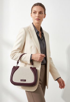 Donna con blazer beige e camicia grigia che tiene una borsa beige e bordeaux Diana & Co Firenze su sfondo bianco uniforme.