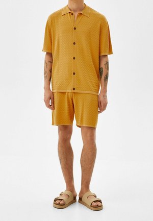 Man draagt een mosterdgele textuur korte mouwen overhemd met knopen en bijpassende korte broek, met beige sandalen met dubbele band, staand.