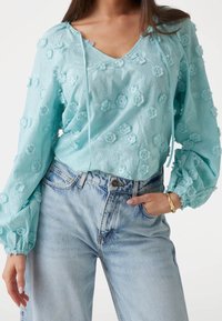 Haut bleu clair avec broderies florales, décolleté en V et détails à cordon. Manches bouffantes, associé à un jean bleu taille haute.