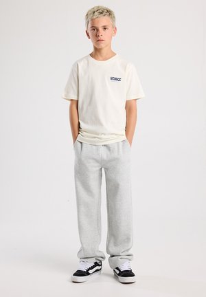 T-shirt col rond en blanc cassé avec "VENICE" imprimé en bleu, associé à un pantalon de survêtement gris clair et des baskets Vans noires. Design simple dans l'ensemble.
