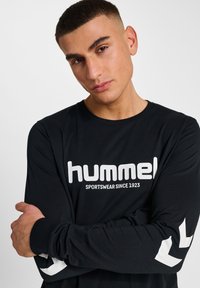 Langärmliges schwarzes T-Shirt mit weißem "hummel"-Logo und Aufschrift. Verfügt über weiße Pfeilakzente an den Ärmeln und besteht aus einem glatten, leichten Material.