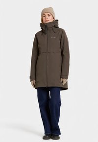 Ruda neperšlampama parka su aukštu apykakliu, reguliuojama kapišonu, priekinėmis užtrauktukais ir šoninėmis kišenėmis, derinama su tamsaus džinso kelnėmis ir smėlio spalvos megzta kepure.