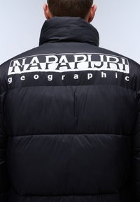 Svart puffjacka med hög krage, med en framträdande vit "NAPAPIJRI geographic" logotyp på baksidan. Slät, vadderad textur.