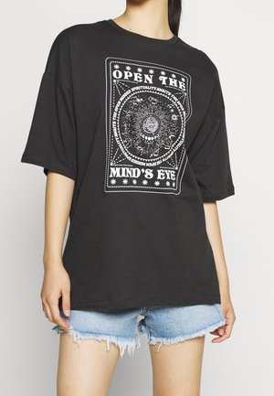 Personne portant un T-shirt noir oversize avec un graphisme mystique blanc représentant le soleil et la lune ainsi que le texte "OPEN THE MIND'S EYE", associé à un short en denim effiloché.