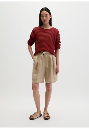 Femme portant un pull rouge texturé à manches longues, un short beige taille haute avec ceinture à nouer et des sandales beiges à boucle, debout les mains dans les poches.