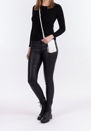 Haut noir à manches longues et pantalon en cuir noir ajusté. Sac bandoulière blanc avec détail zippé. Bottes de combat noires à lacets. Fond uni.