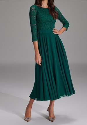 Femme en robe en dentelle bleu sarcelle avec manches trois-quarts et jupe fluide, portant des talons transparents ornés de strass.