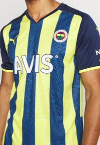 Żółto-granatowa sportowa koszulka w paski z abstrakcyjnymi wzorami, logo klubu sportowego na piersi oraz wyraźnym logotypem "AVIS".