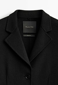 Manteau en laine noire avec col cranté, fermeture par un bouton unique, et tissu lisse texturé. Étiquette visible à l'intérieur avec le nom de la marque.