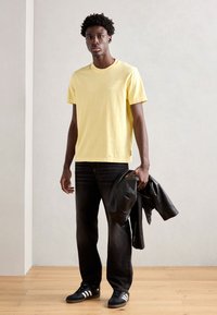 Yellow short-sleeve t-shirt, relaxed fit; black jeans; holding a black leather jacket; black sneakers with white stripes; wooden floor background.