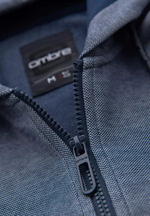 Chaqueta azul con cremallera, que presenta un tejido texturizado, cuello en contraste y etiqueta de logo. Detalle de cremallera visible y costuras.