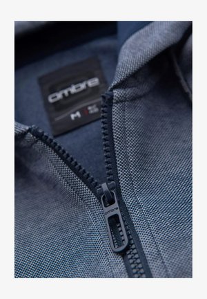 Blauwe jack met rits van textuurstof, met contrasterende kraag en logolabel. Zichtbare ritsdetails en stiksels.