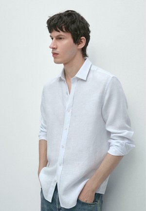 Junger Mann mit dunklem Haar, der ein weißes Hemd mit Knöpfen und blaue Jeans trägt, steht mit den Händen in den Taschen vor einer schlichten weißen Wand.