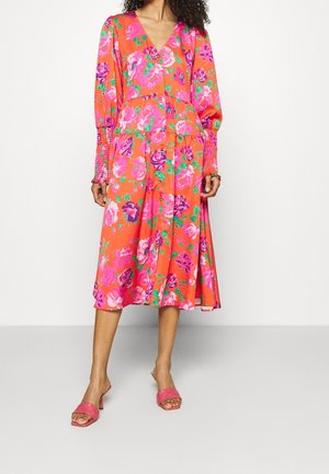 Kvinde iført en klart orange midi-kjole med pink og lilla blomsterprint, lange puffede ærmer og pink hælede sandaler.