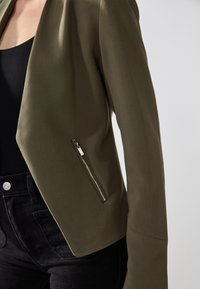 Olivgrön kort blazer i ett slätt tyg, med en silverfärgad dragkedjeficka på vänster sida och en strukturerad, öppen frontdesign.