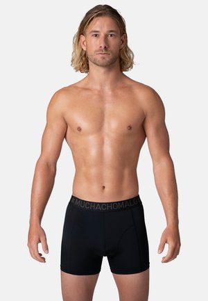 Zwarte boxershorts met een logo op de tailleband, slim fit ontwerp, gemaakt van gladde stof. Model staat met de armen ontspannen langs de zijden, en toont de pasvorm van het product.