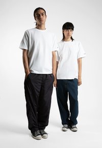 Two individuals wear plain white cotton t-shirts. One pairs it with black nylon pants; the other with blue denim jeans and black sneakers.